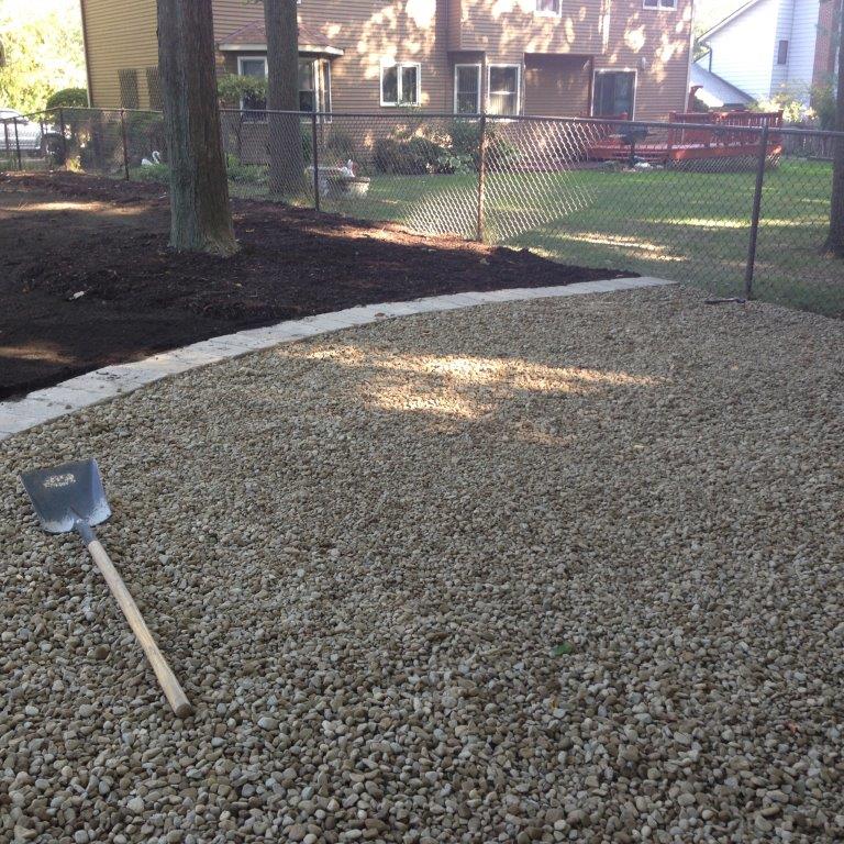 Shovel on a gravel yard beside a tree and chain-link fence in a backyard setting.
