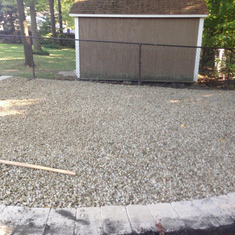 Gravel yard beside a tan shed with chain-link fence and stone border in the foreground