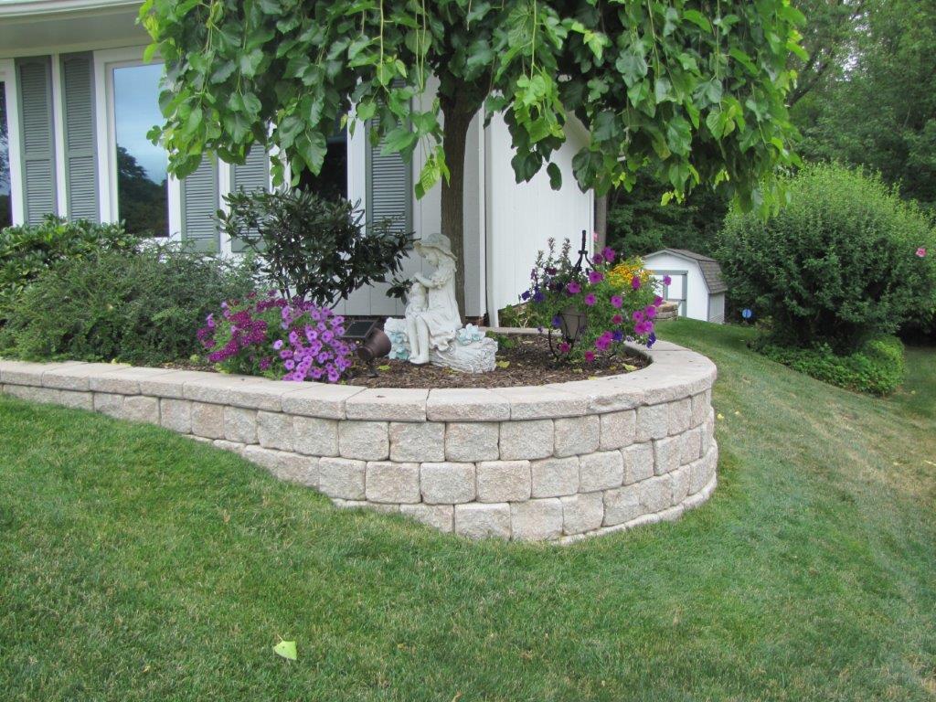 Front yard garden bed with purple flowers around a tree beside a house, bordered by tan stone blocks