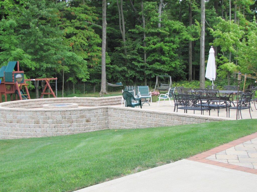 Backyard patio with stone wall, outdoor seating, umbrella, and green lawn by a wooded area