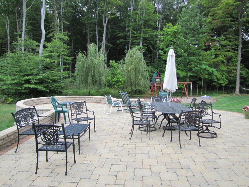 Patio with metal chairs, tables, and a closed umbrella beside a green wooded yard