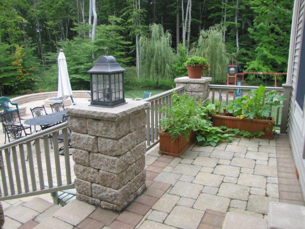 Patio with stone pavers, lantern-topped pillars, potted plants, and a view of a pool and wooded yard