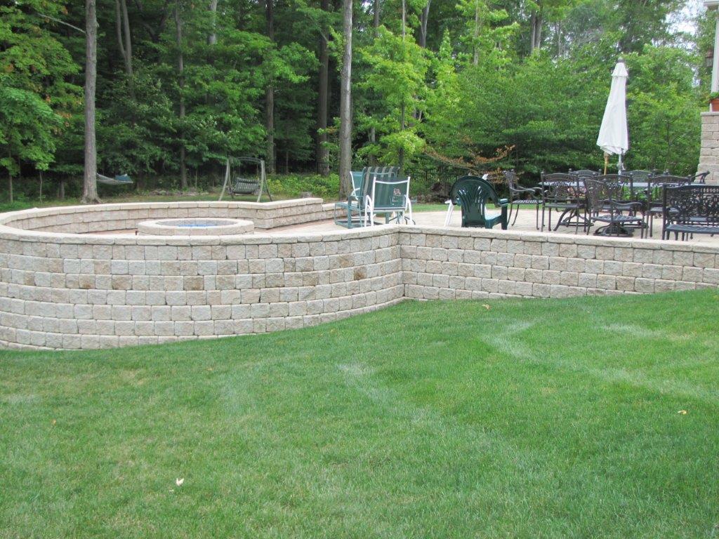 Stone patio and low retaining wall beside a green lawn and wooded backyard seating area.