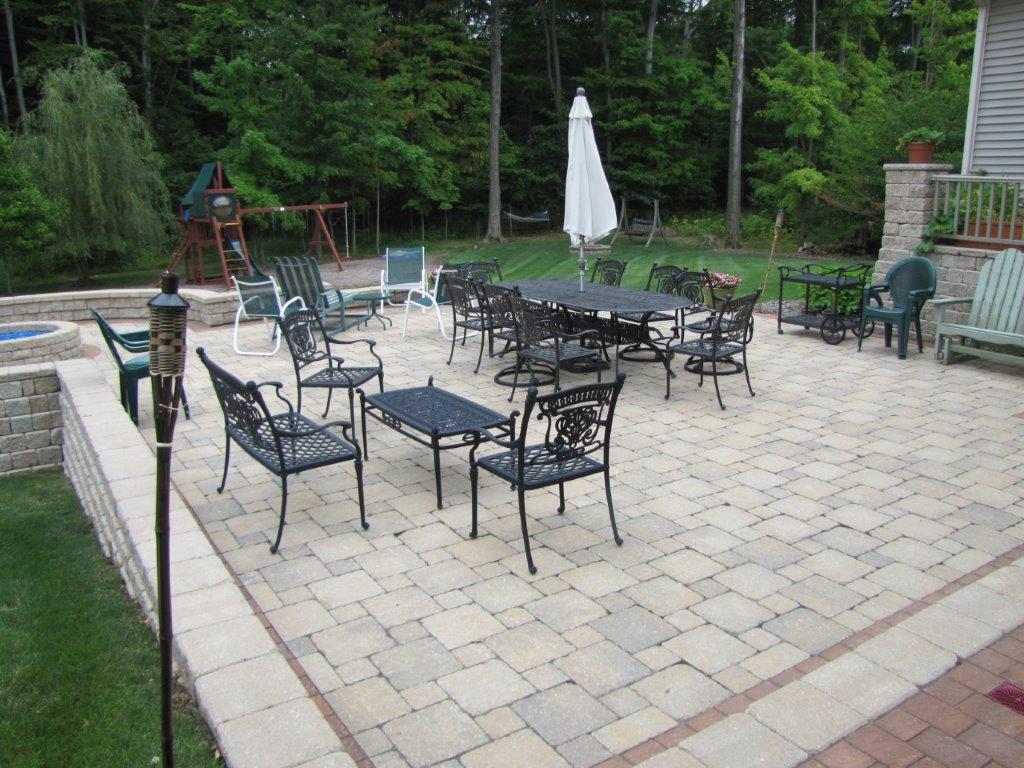 Outdoor patio with black metal tables and chairs, umbrella, and grill beside a wooded backyard area