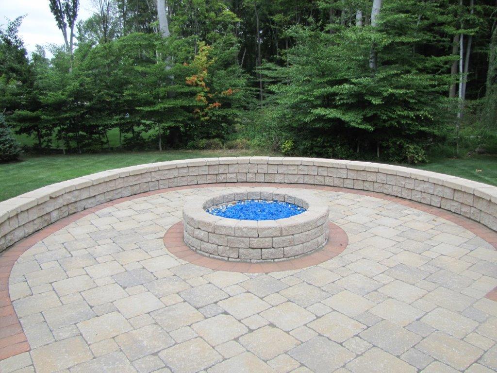 Circular stone fire pit in a paved patio, surrounded by a curved bench and green trees.