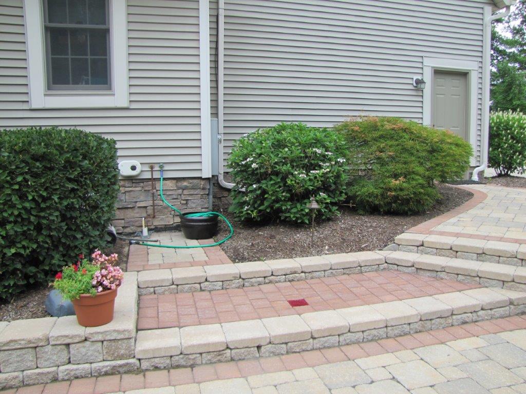 Front yard garden with stone steps, potted flowers, shrubs, and a hose by a house siding.