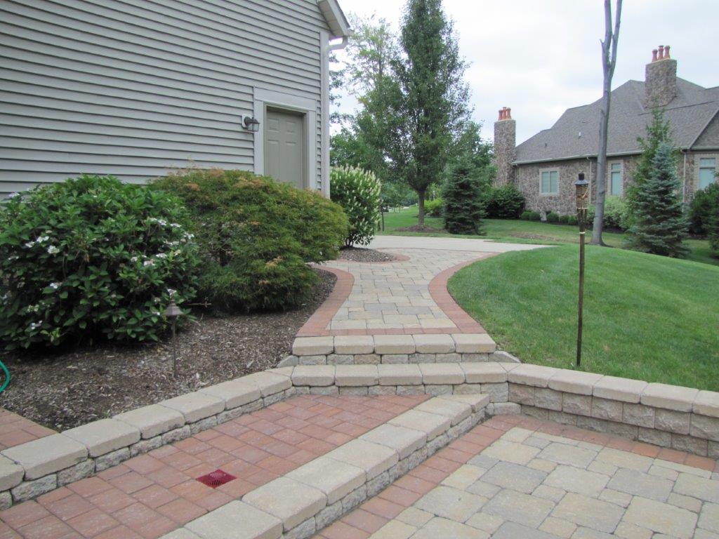 Brick walkway and patio leading past a house and lawn in a suburban yard.
