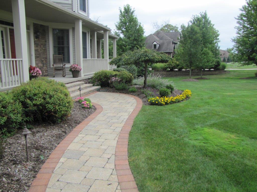 Curved stone walkway beside a house with porch, shrubs, flowers, and a green lawn.