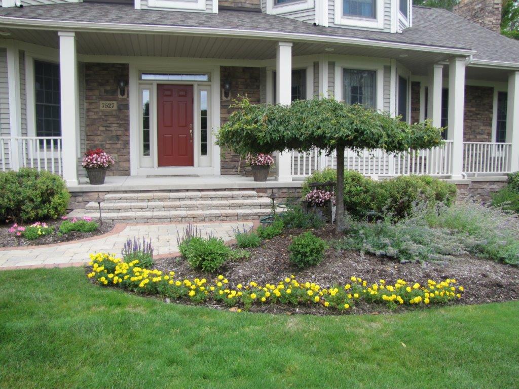 Front porch of a gray house with red door, white columns, and a flower-filled garden border.