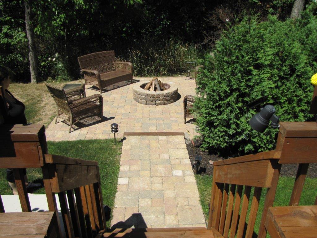 Backyard patio with stone fire pit, chairs, and a paver path viewed from wooden stairs