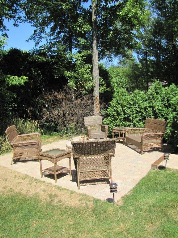 Patio seating area with wicker chairs around a small table under trees in a sunny garden