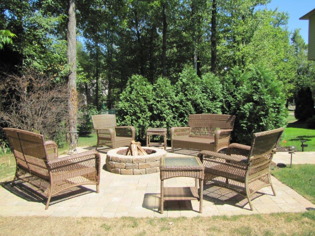 Wicker outdoor seating around a stone fire pit on a sunny patio surrounded by trees and shrubs