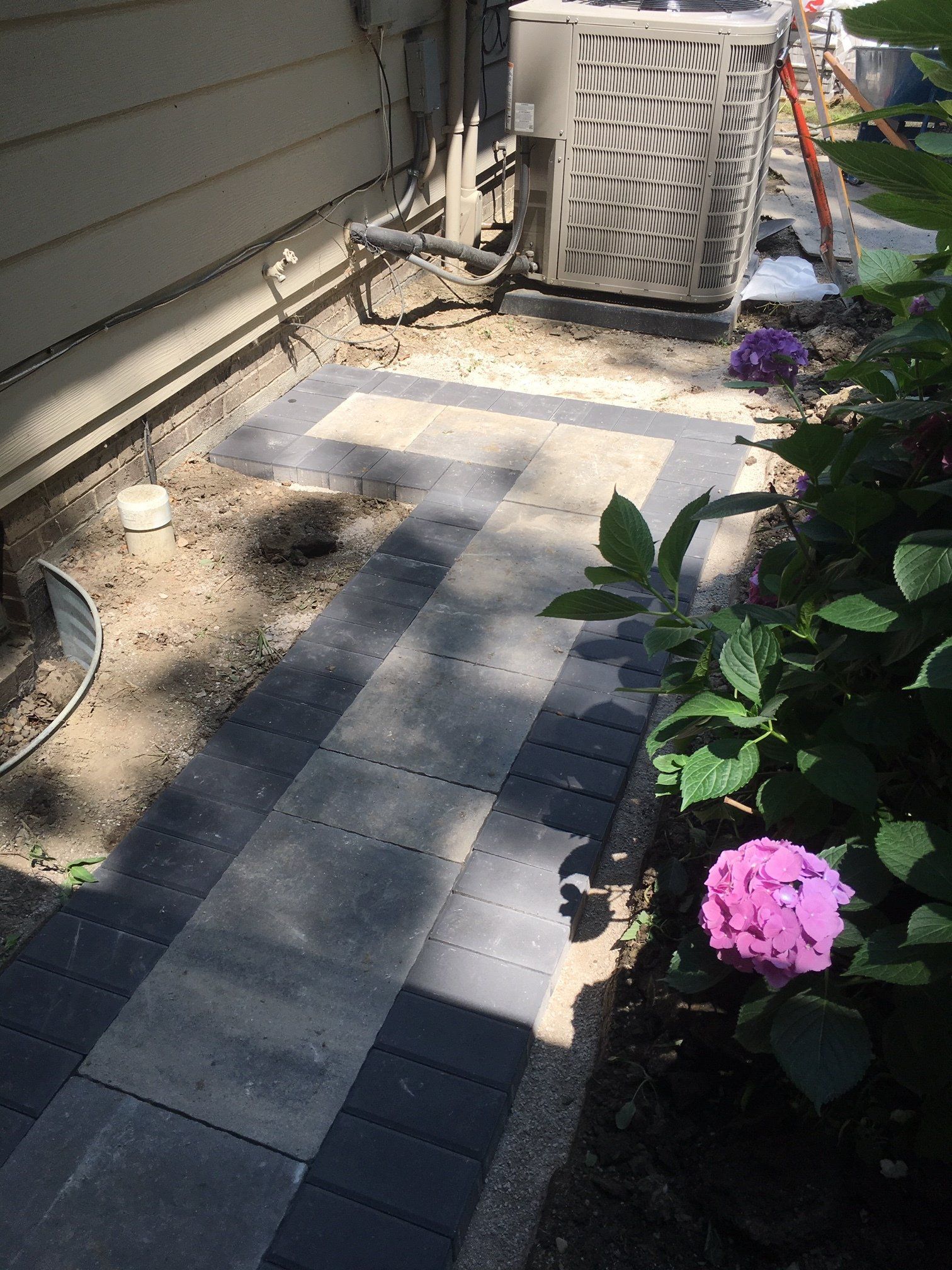 Narrow garden path beside house with air conditioner, flowers, and shadows on pavers