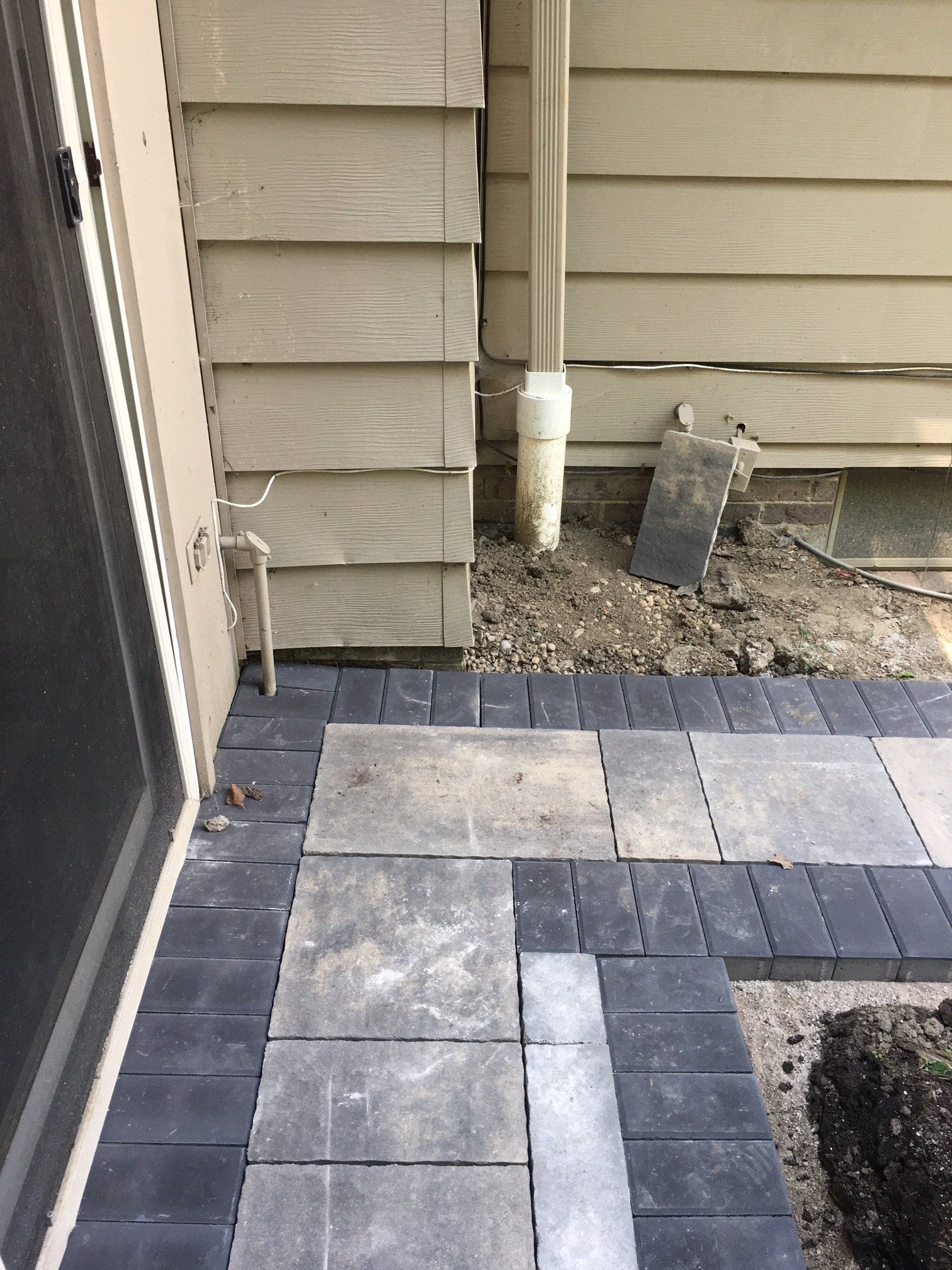 Stone paver patio beside a house wall, with dark border tiles and a downspout on gravel.