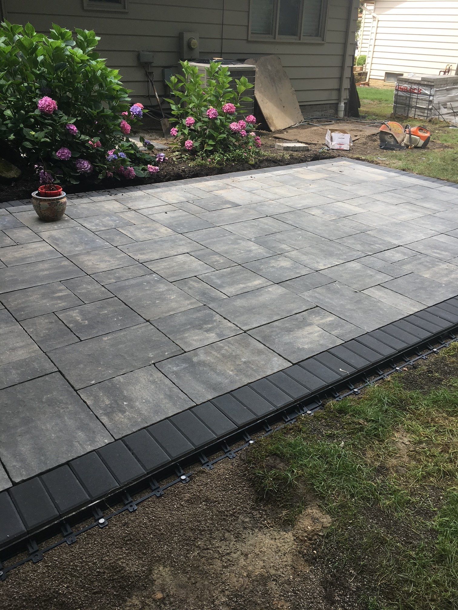 Backyard patio with large gray pavers, dark border edging, and flowering shrubs along the house