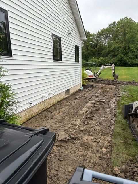 Dug-out dirt strip beside white house, with excavator in yard and dark bins in foreground