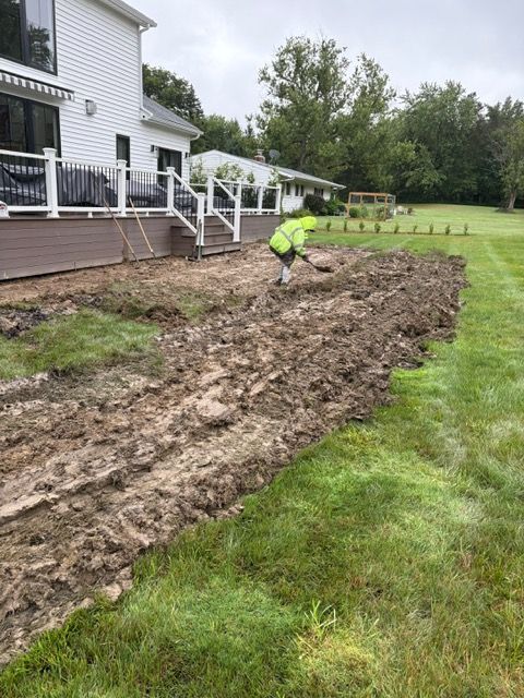 Worker in neon jacket leveling muddy soil beside a house with white porch railings.