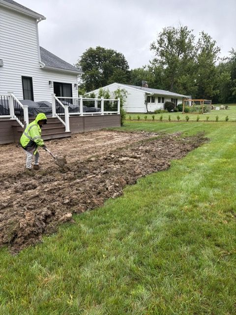 Person in neon jacket working in a muddy yard beside a white house and deck.