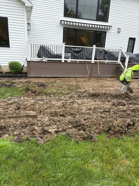 Muddy backyard beside a deck, with a person in a bright jacket working near the grass edge.