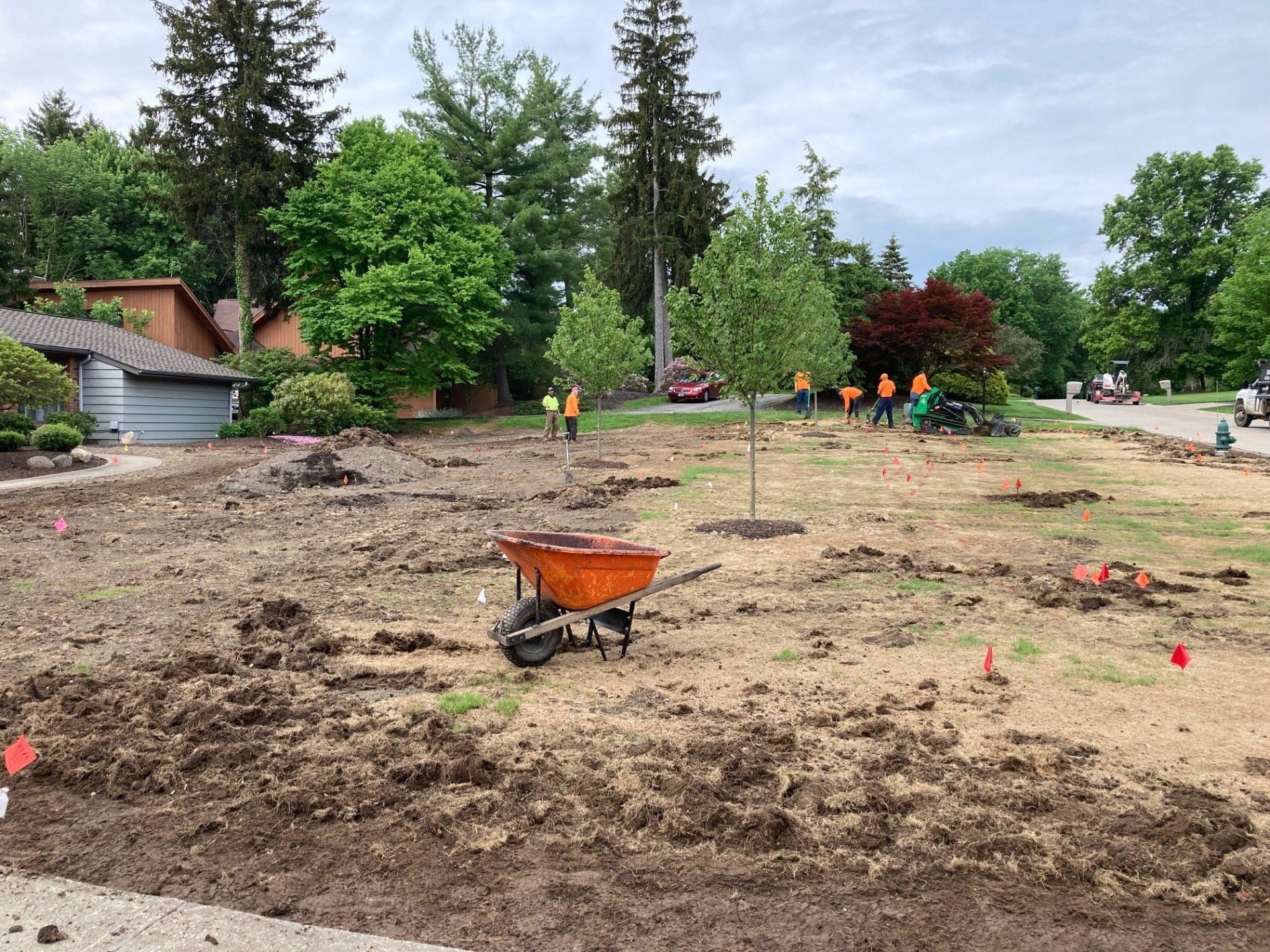 Construction site with dirt lot, orange wheelbarrow, and workers in orange vests near trees