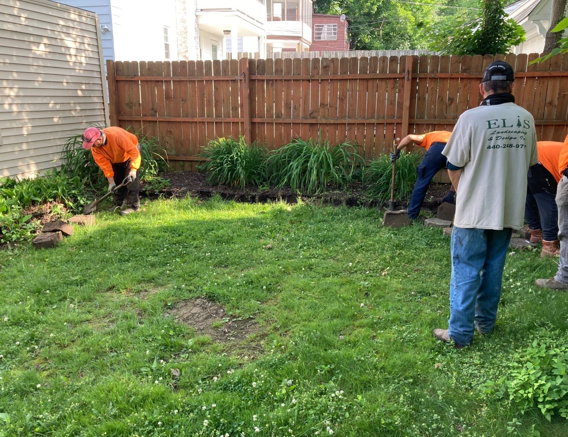 Workers trimming a backyard lawn and garden beds by a wooden fence