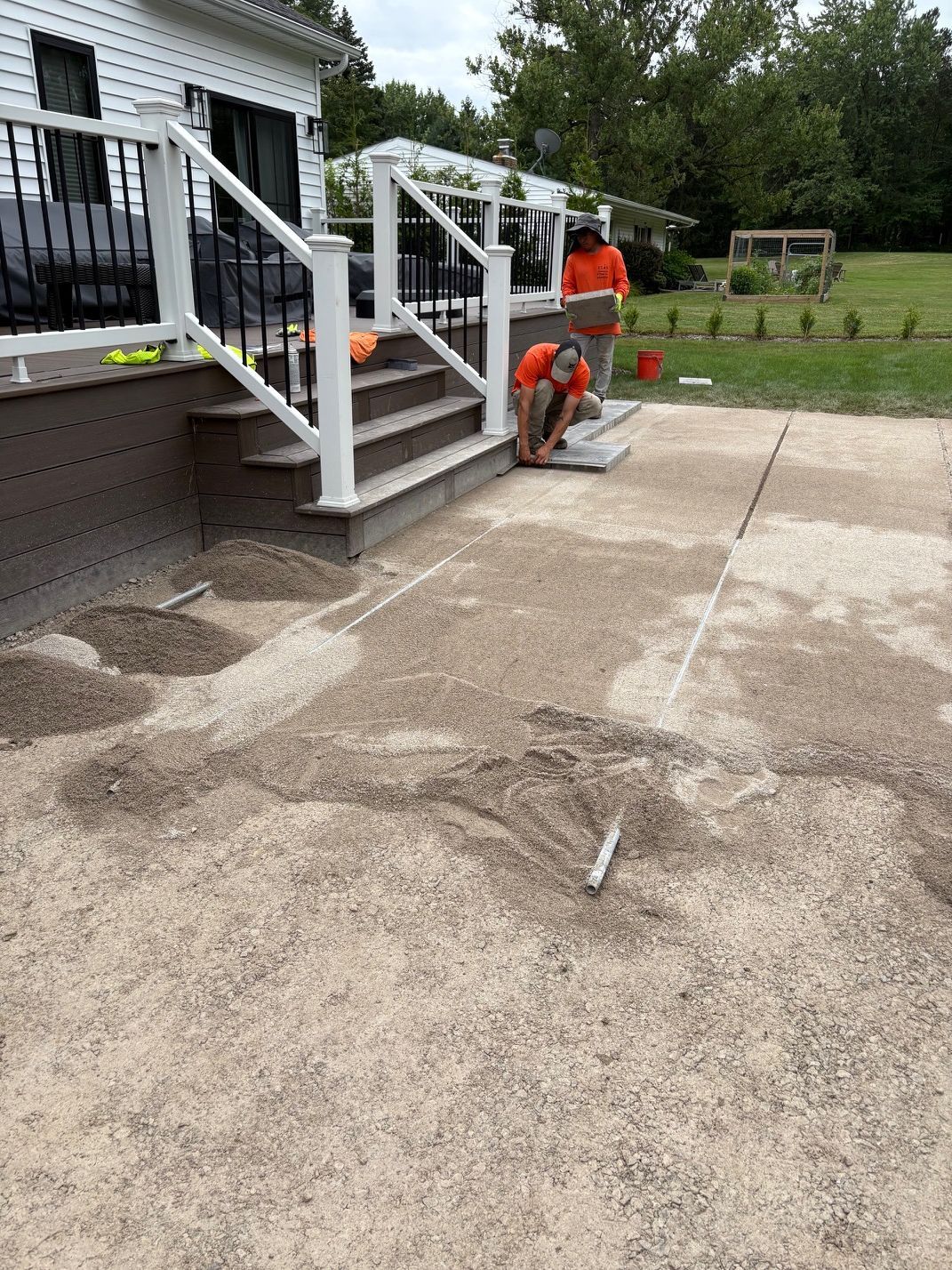 Workers repairing a concrete patio and white stairs beside a house, with one worker kneeling near tools.