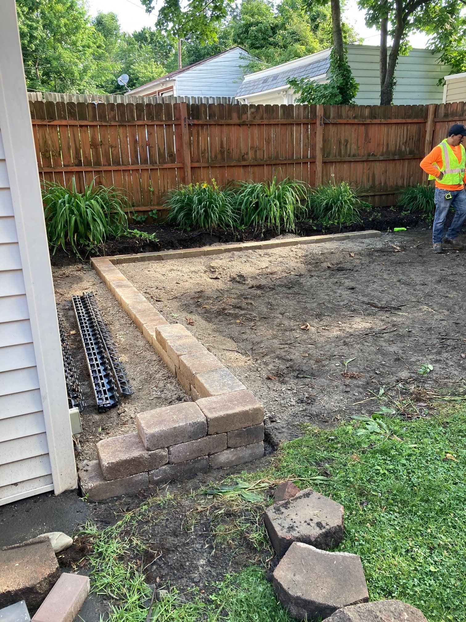 Backyard construction site with raised edging, dirt lot, fence, and worker in orange vest