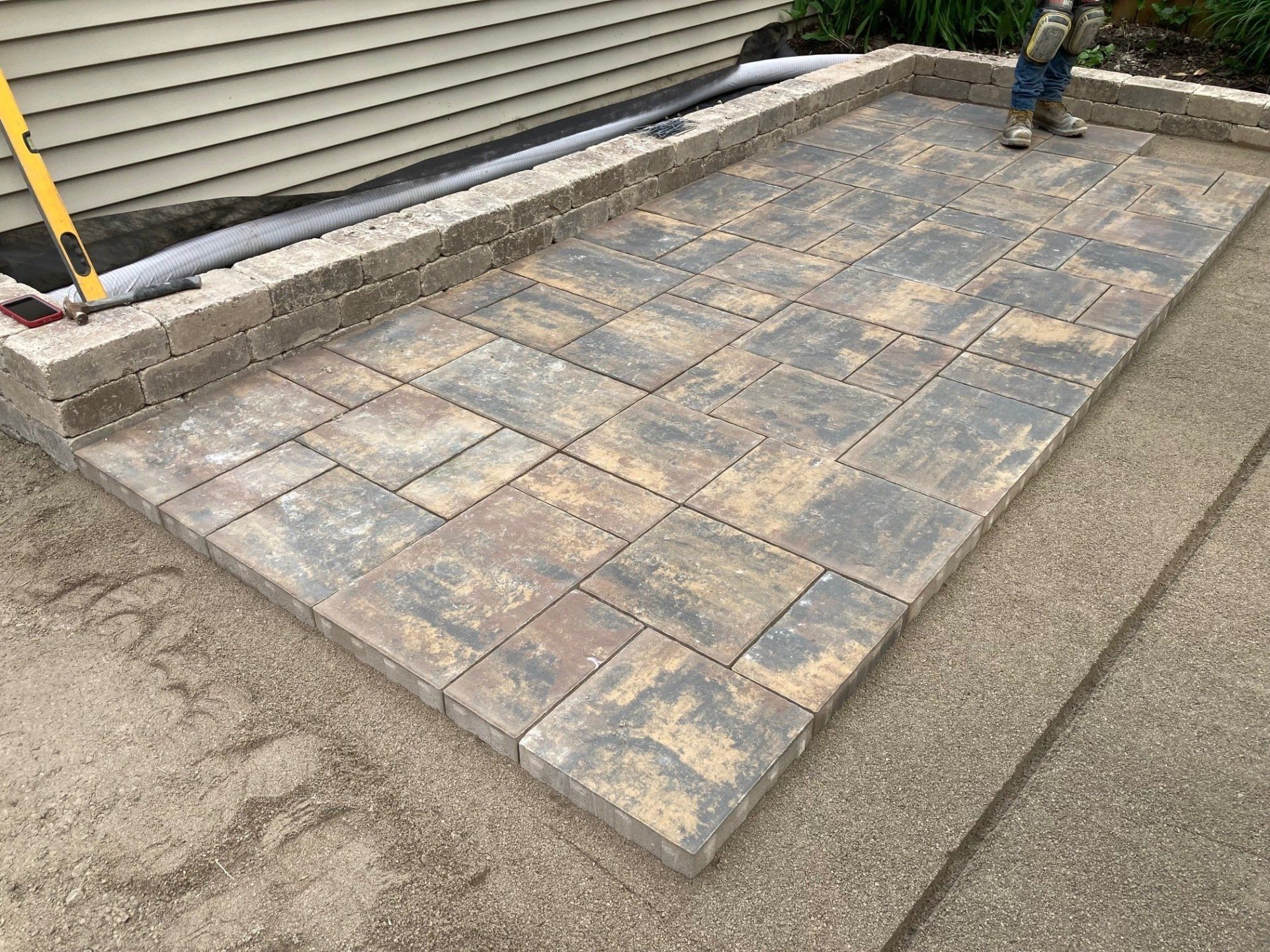 Paver patio under construction beside a house, with workers and tools visible.