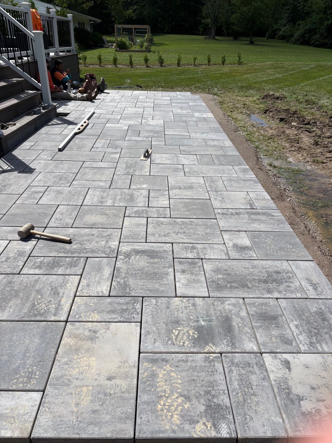 Stone patio under construction beside a grassy yard, with tools and a staircase on the left.