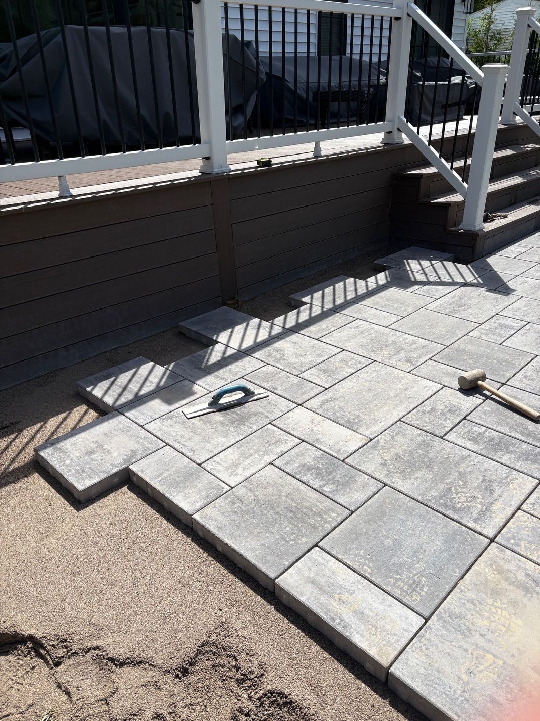 Patio pavers beside a railing, with a step, drain cover, and part of a gravel path in sunlight