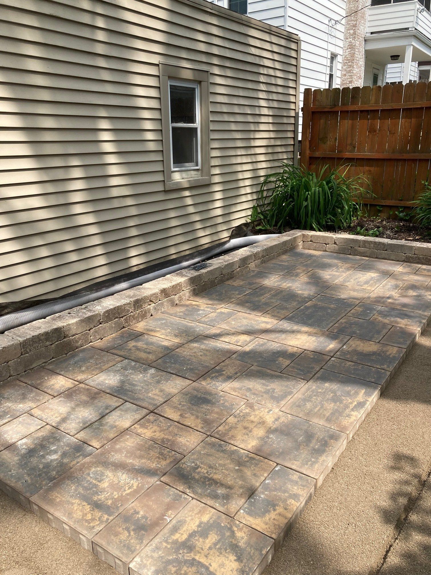 Backyard patio with paver stones beside a house and wooden fence, near a small garden bed.