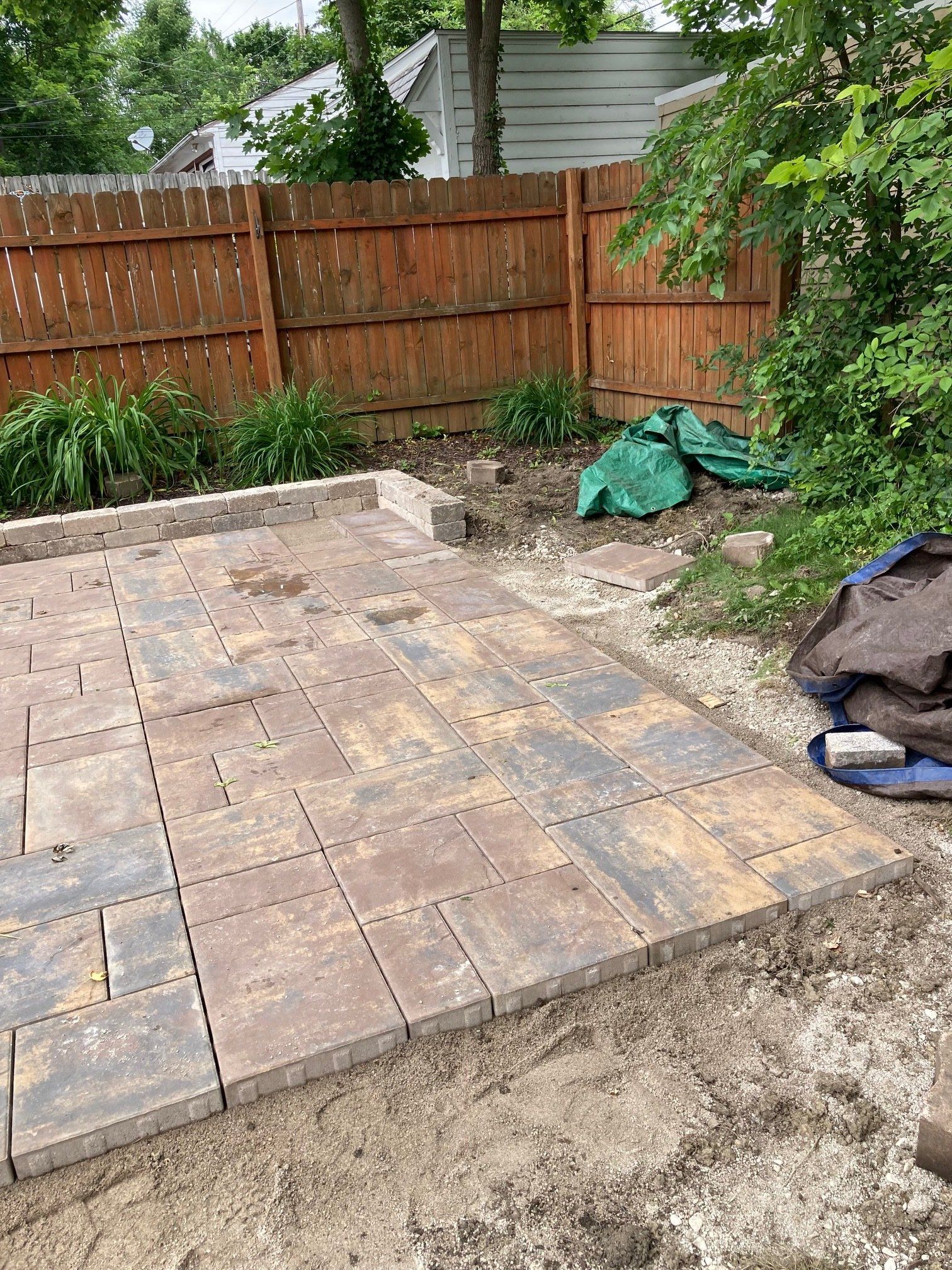 Backyard patio of square pavers beside a wooden fence, with plants and covered yard items.