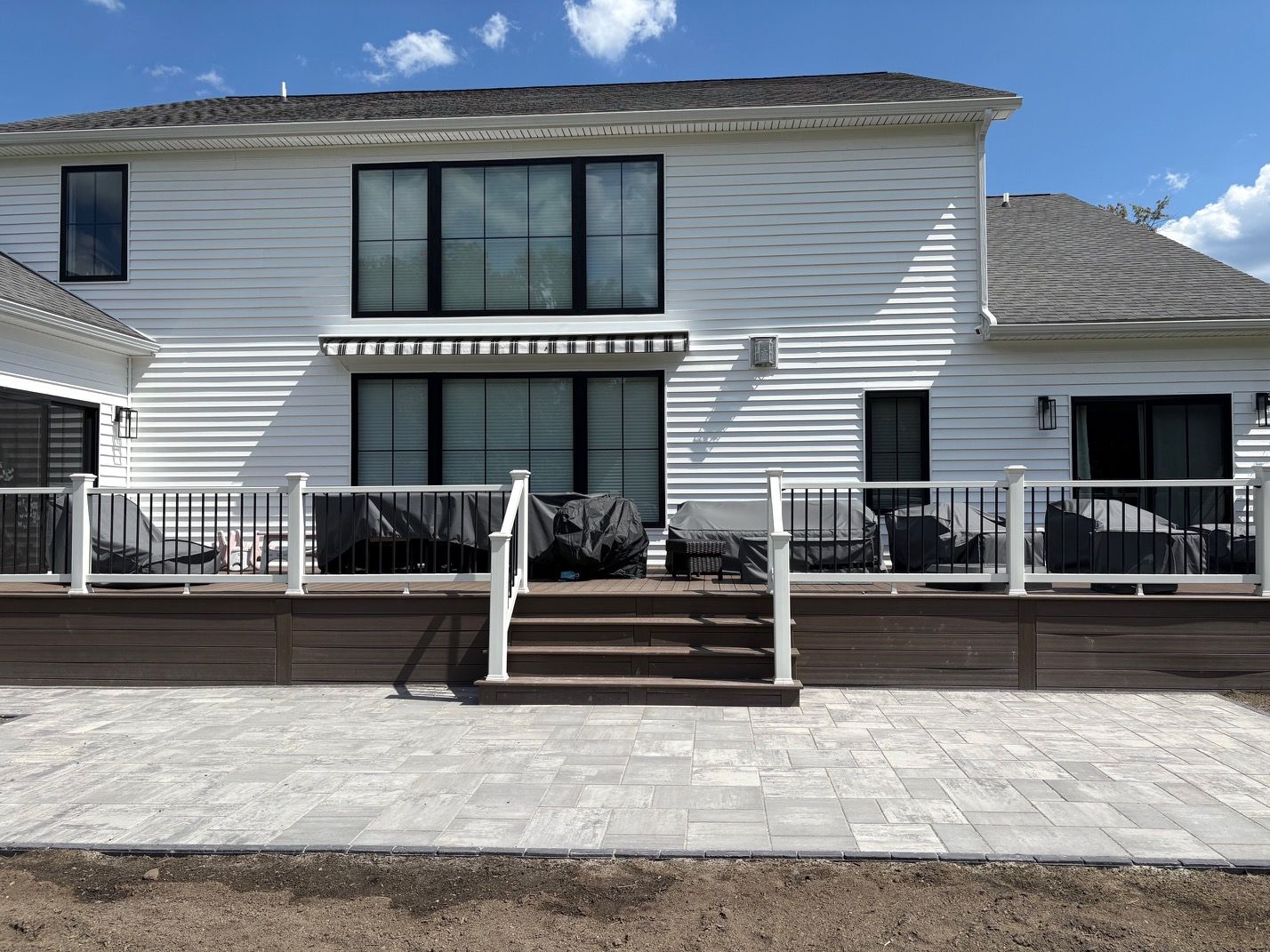 Front view of a white two-story house with a deck, stairs, and a paved patio under a blue sky