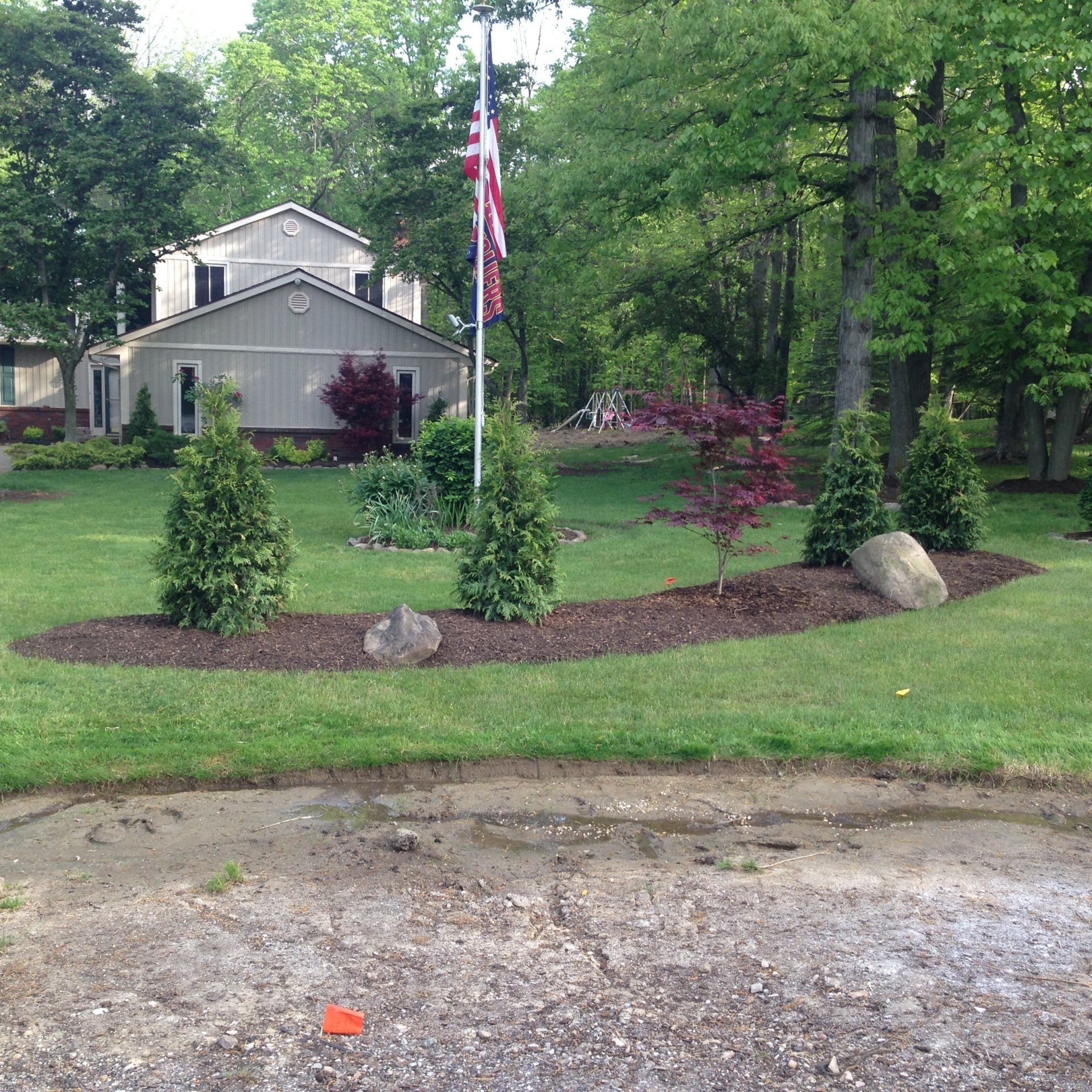 Front yard landscaping with flagpole, shrubs, mulch beds, and a house in the background, beside a gravel driveway