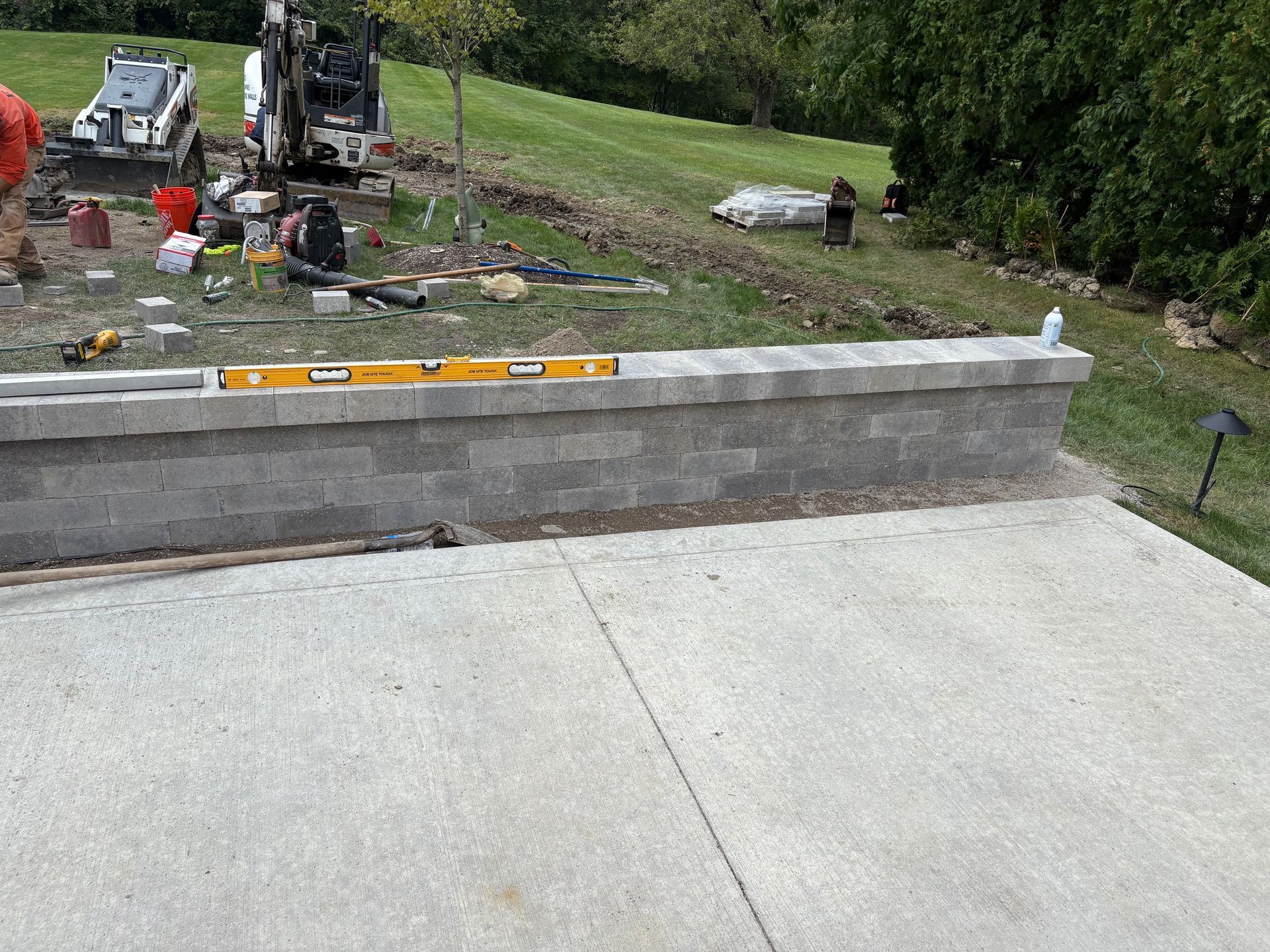 Concrete patio with a low cinder block wall; construction equipment and materials in a grassy yard beyond.