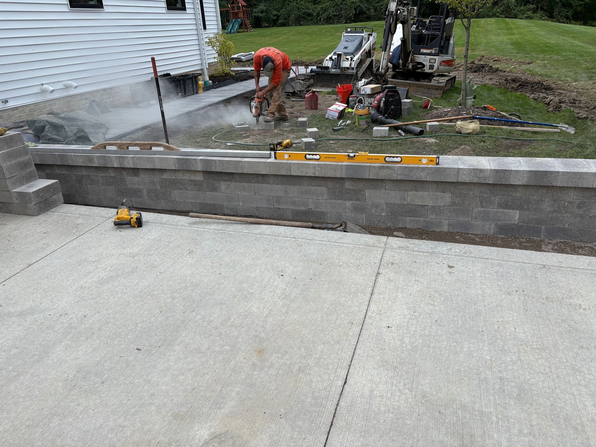 Workers building a gray concrete retaining wall and driveway beside a house, with tools and equipment nearby