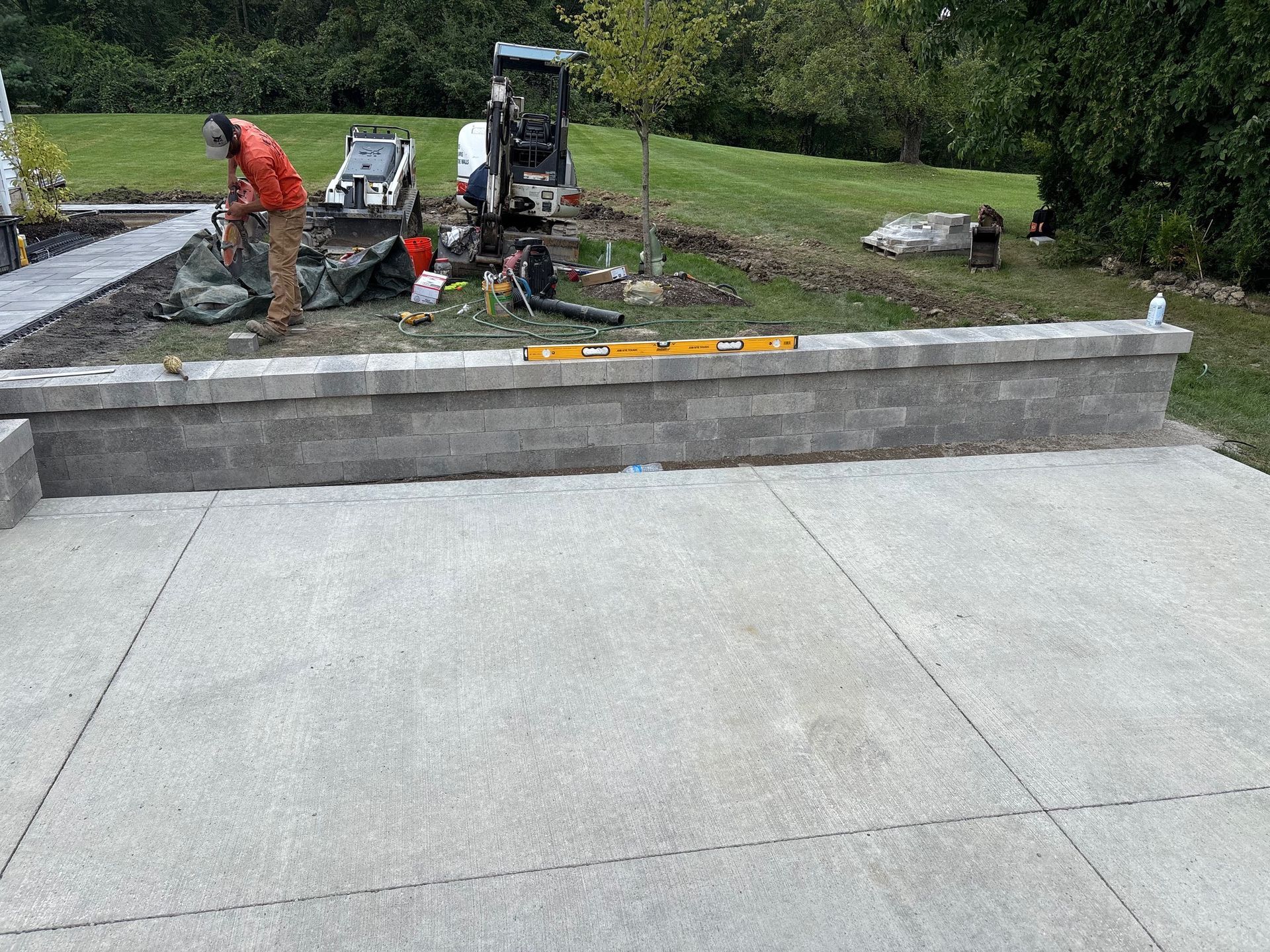 Concrete patio overlooking a landscaped yard, with workers and equipment building a low retaining wall.