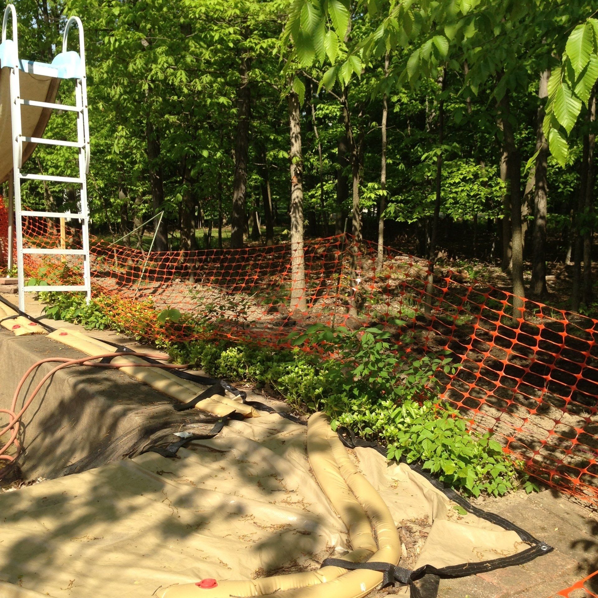 Backyard under construction with orange safety fencing, tarp-covered ground, and a white ladder by trees