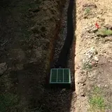 Narrow dirt trench with a green drainage grate at the bottom