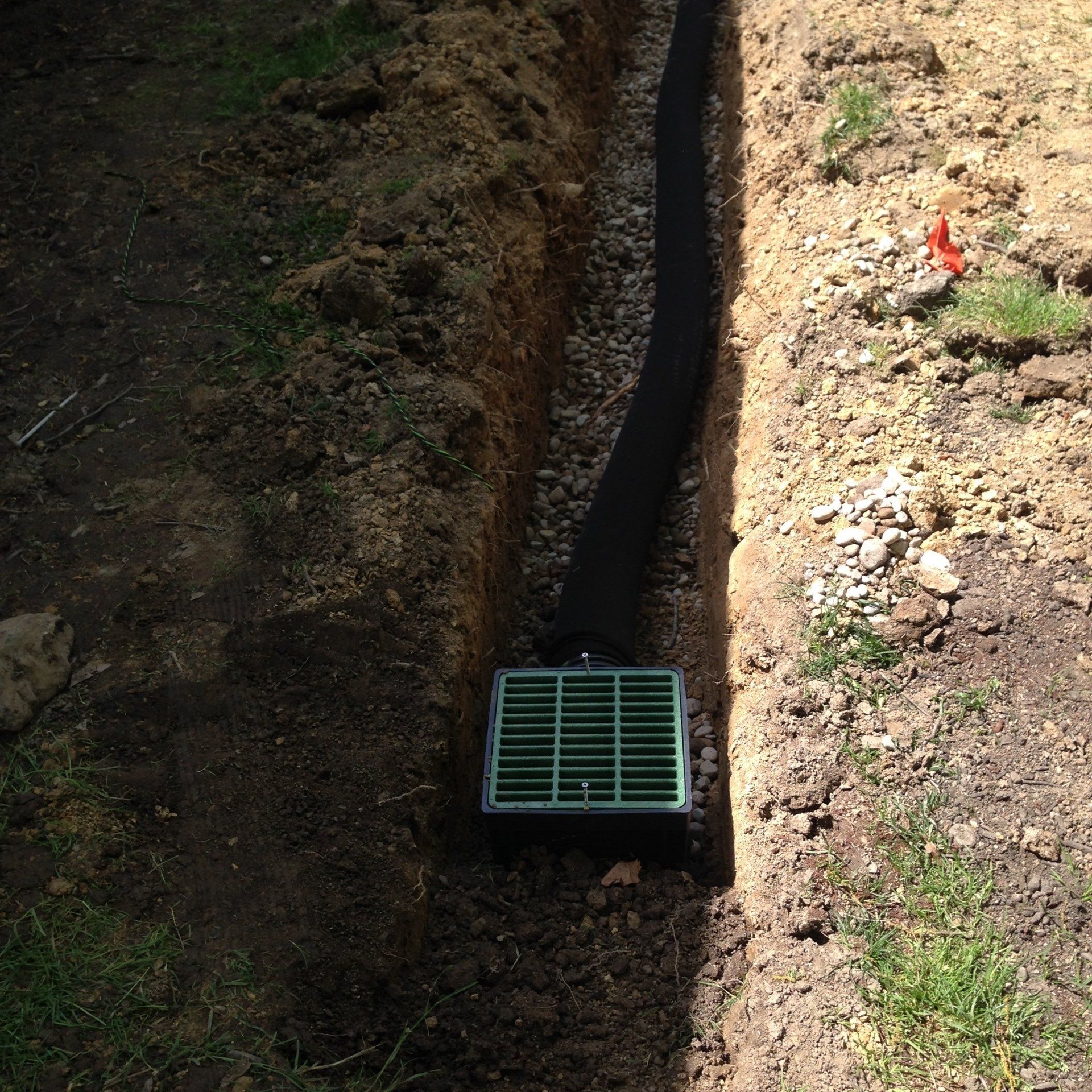 Narrow dirt trench with a green drain grate at the bottom, bordered by grass and sunlight.
