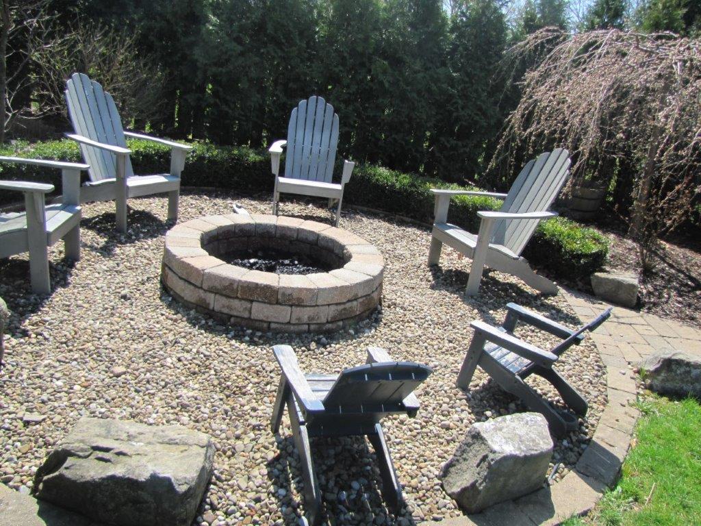 Backyard fire pit with six wooden chairs arranged around it on a gravel patio.