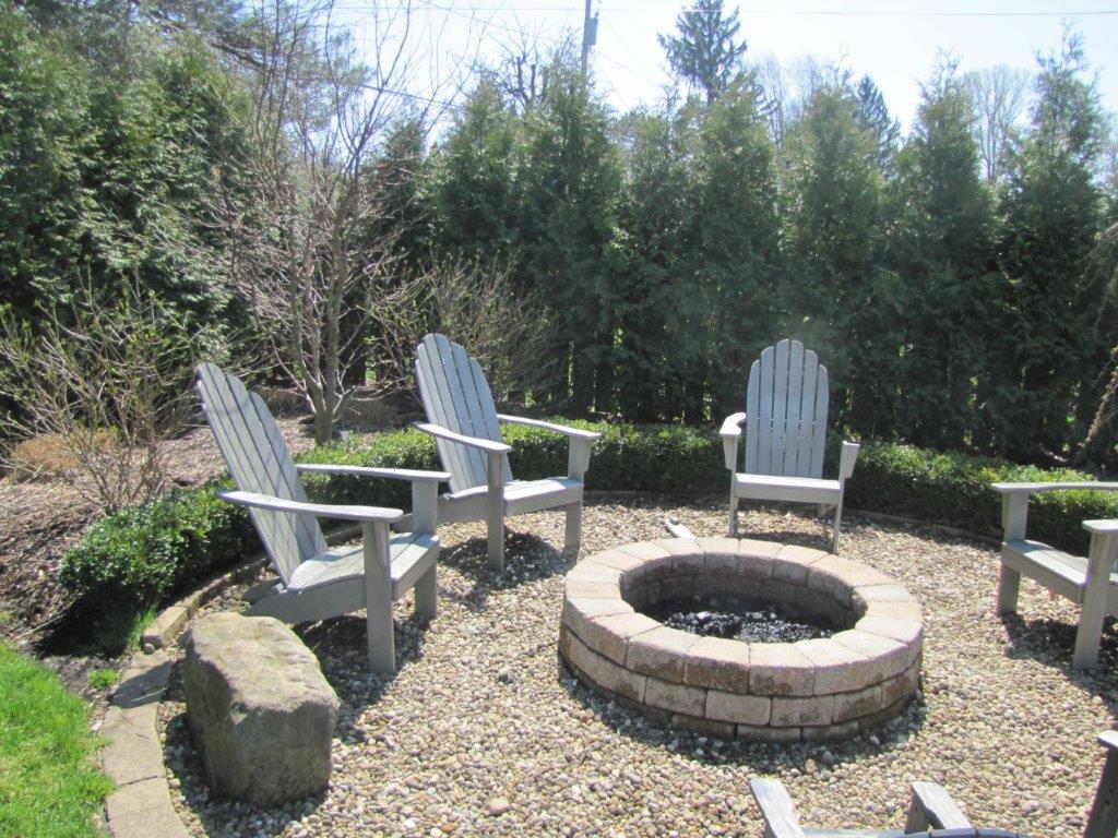 Three white chairs around a stone fire pit in a gravel backyard surrounded by trees