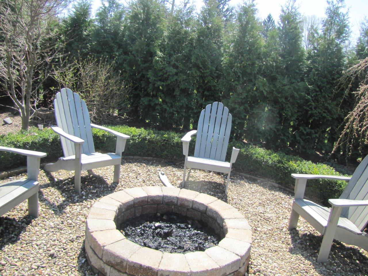 Backyard fire pit surrounded by white Adirondack chairs on a gravel patio with trees behind
