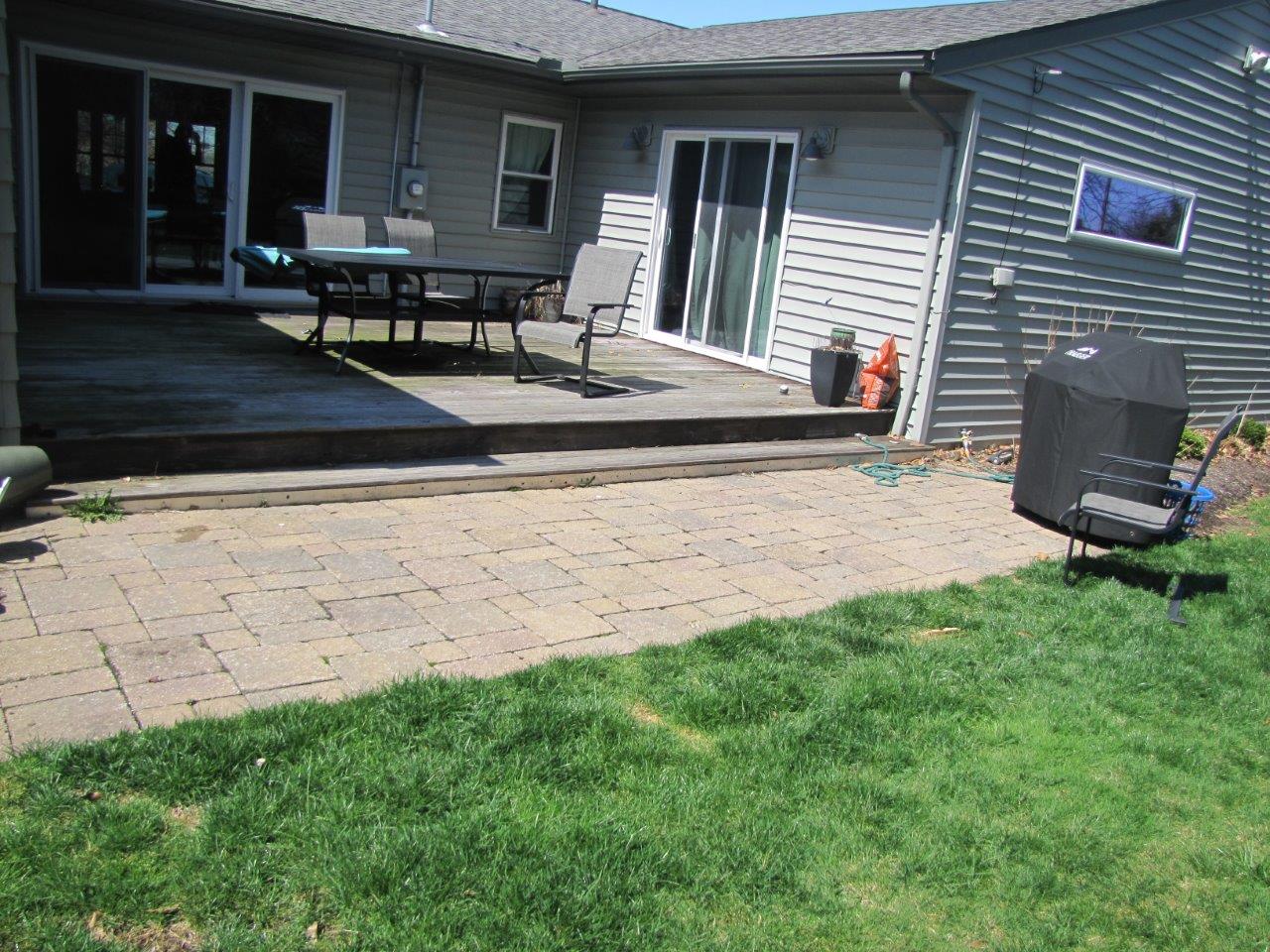 Backyard patio with stone pavers, outdoor table, grill, and sliding glass doors beside a grassy lawn