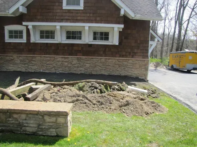 Stone house with a dirt pile and broken branches in the yard beside a road.