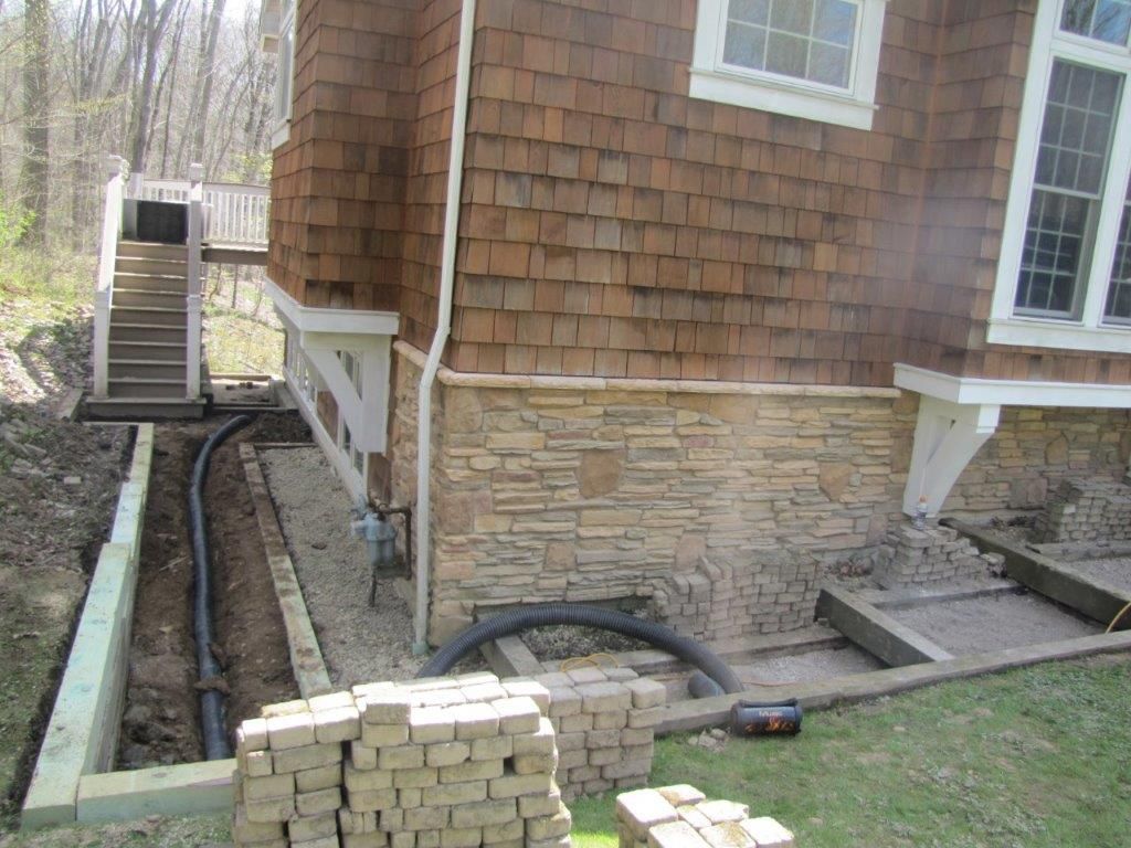 Basement wall excavation with black drainage pipe and stone veneer house exterior