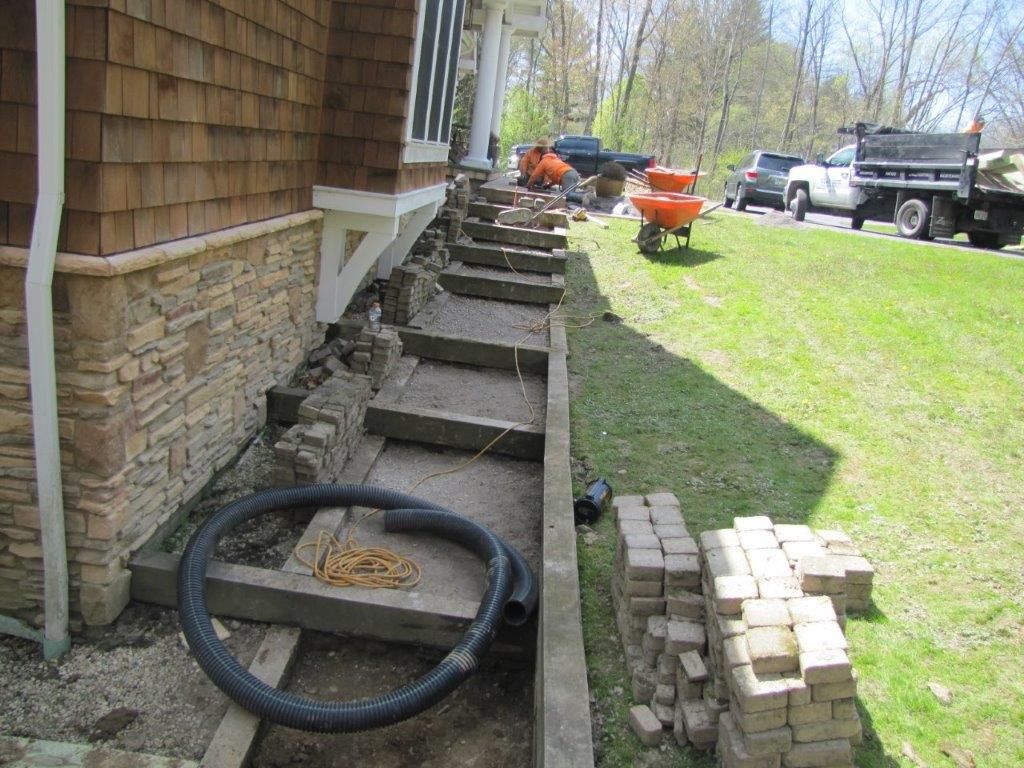 Trench along house foundation with pipes, stones, and tools on a residential lawn during outdoor work