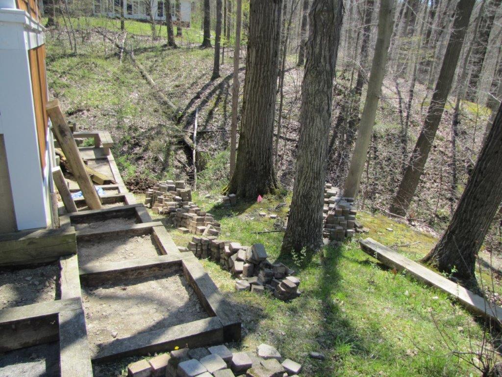 Stairs beside a house lead down a wooded hillside with stone edging and scattered steps.