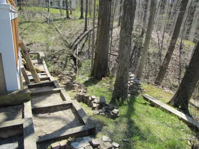 Wooden steps and stone path beside a house on a wooded hillside with trees and grass