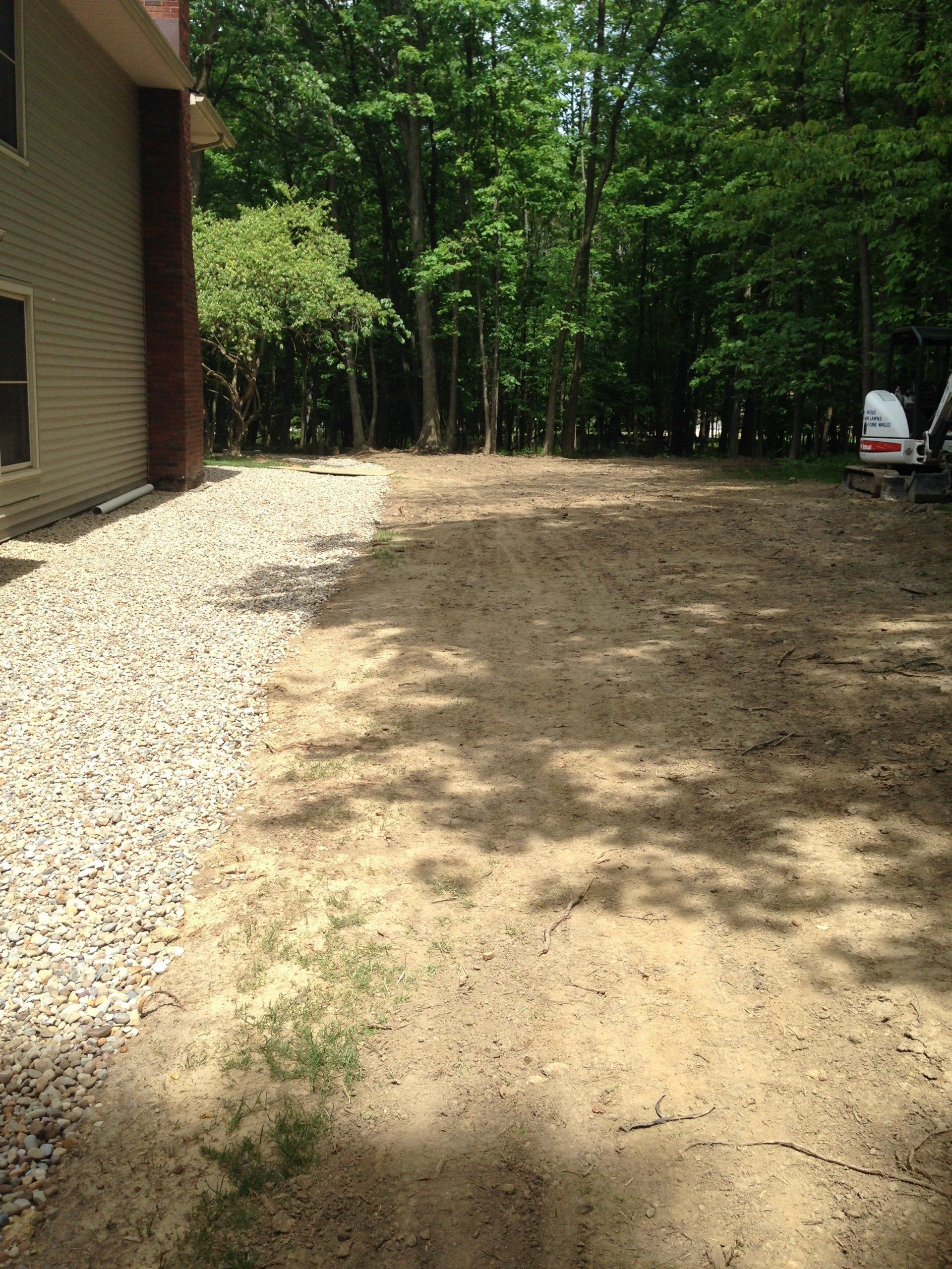 Gravel path beside a house leading to a wooded backyard, with a dirt area and a parked trailer in the distance